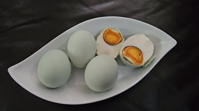 egg in a bowl,Five Chinese salted eggs rest on a leaf-shaped plate. Three eggs remain whole, showcasing their pale blue shells. One egg is cut open, revealing a firm, golden yolk. Another egg is halve