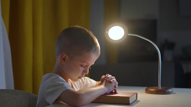Boy praying quietly with hands folded on bible. Faith visible in boy posture. Hope shines as boy praying under lamp. Boy praying shows worship and trust. Peaceful child moment of faith and hope