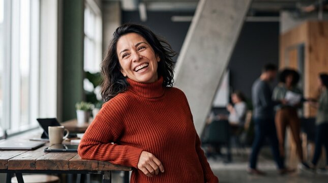 A cheerful middle aged Hispanic businesswoman in a rust sweater laughing in a modern open plan office environment