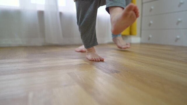 Little child steps carefully on hardwood floor. Barefoot toddler tries walk beside adult. Baby practicing balance step indoors. Boy leg moves forward during first walk. Child learning to walk at home.
