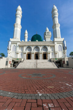 White mosque stands tall under bright blue sky. Green dome gleams above arched entrances and twin minarets. Broad stone steps lead to grand doors flanked by decorative arches