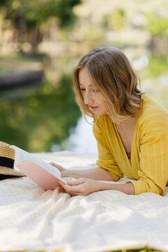 Woman reading book by lake on picnic blanket