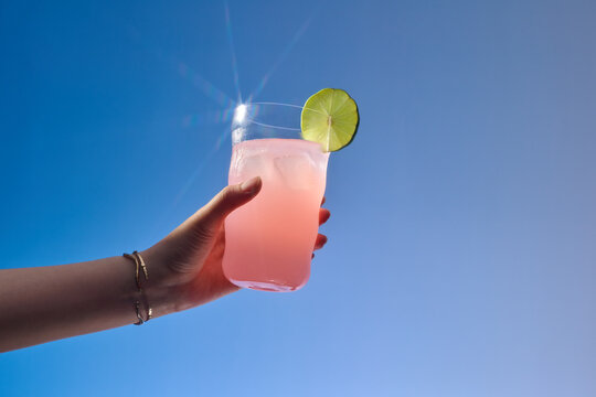 Hand holding up a pink lemonade with a lime slice against blue sky