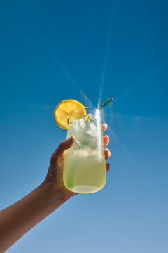 A hand holds a glass of refreshing lemonade over blue sky