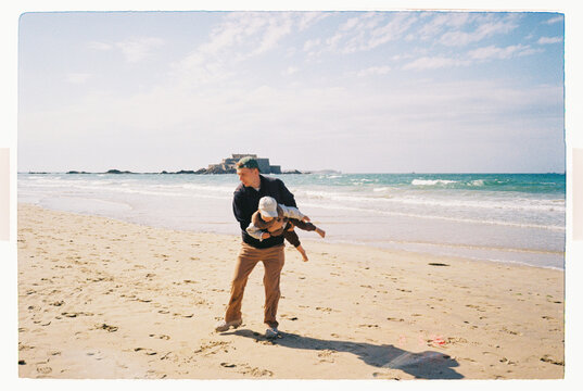 Dad spinning toddler around on a sandy beach by the sea