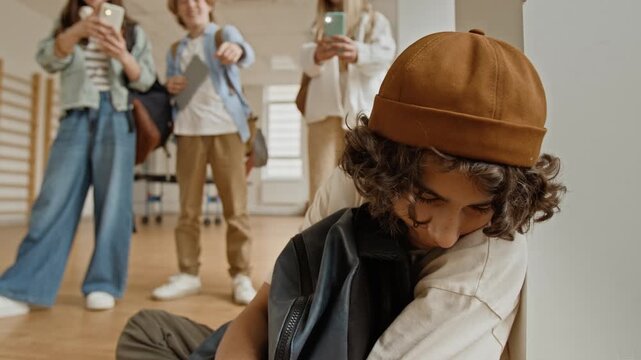 Caucasian teenager boy sitting on school hallway floor, feeling sad and excluded while classmates pointing fingers and recording him on smartphones. Clip is ideal for anti-bullying campaigns