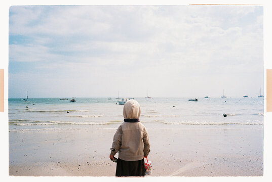 Unrecognizable child holding bucket of seashells watching boats by sea