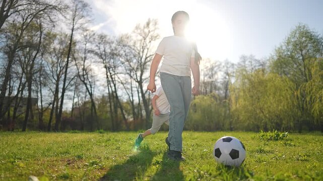 Girl controls football while boy watches. Ball on grass during sunny outdoor day. Children play football together. Girl runs after ball. Child smiles during football play. Ball, football outdoor joy.