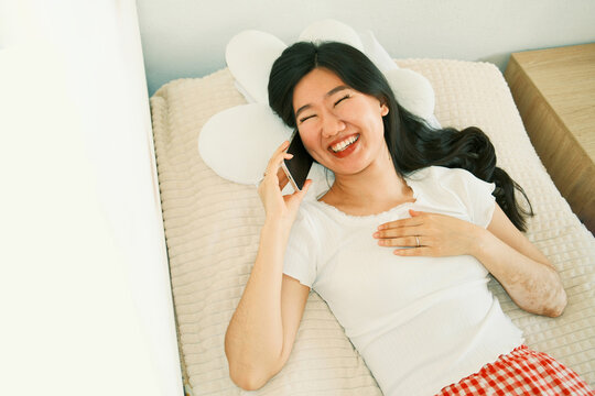 Overview of happy young asian woman talking on smartphone in bed 