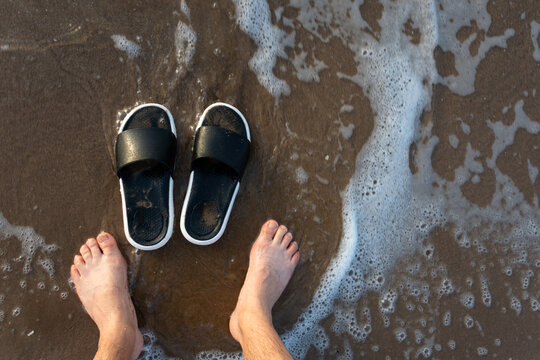 Close up of man's feet standing on beach sand with flip flops by sea