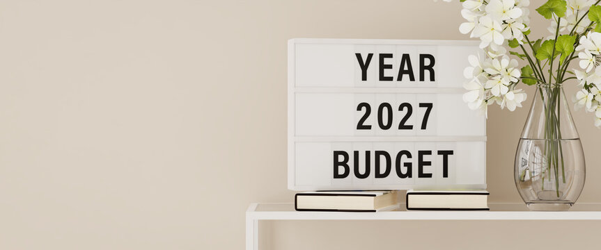 A lightbox displaying the words "Year 2027 Budget" on a shelf with books and plants. Copy space, web banner format.
