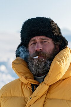 Winter portrait of a man with a frosted beard wearing a black ushanka and yellow down coat smoking outdoors for editorial portrait and cold weather design