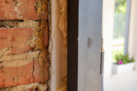 Close-up of the gap between a dark metal door frame and a red brick wall, filled with hardened mounting foam. A metal hinge and plastic plug are visible on the doorjamb during entrance door installati