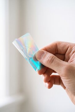 A close-up shot of a person holding an iridescent holographic oral dissolve film strip between fingers against a bright white background