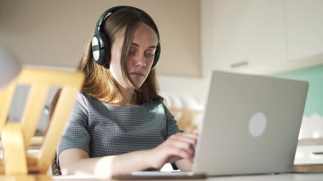 Woman typing on laptop wearing headphones. Remote worker uses computer at home desk. Freelancer works on laptop near kitchen window. Woman with headphone types on notebook computer. Online study.