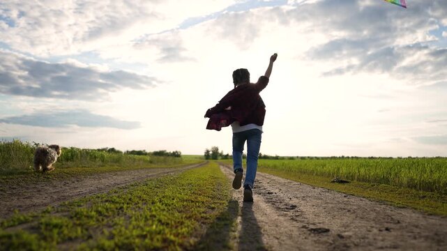 Active girl running fast down dirt road. Happy child playing outside flies large kite. Pet dog chases running girl through green field. Summer sunset brightens open field. Dog loves flying kite.
