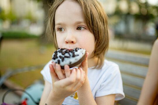 Young boy eating a chocolate donut while sitting on a park bench outdoors. Sweet snack, childhood treat and relaxed leisure during a summer walk.