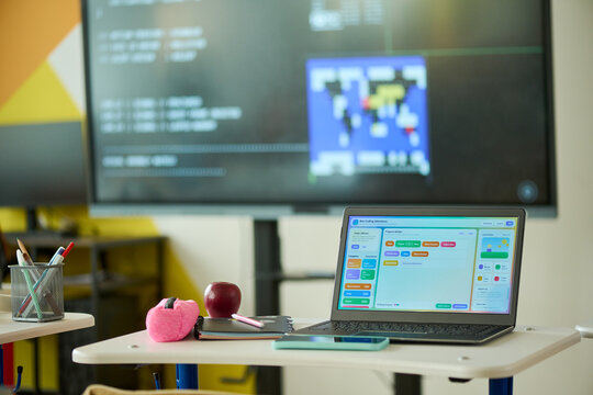 Laptop displaying block coding software on desk in empty classroom with interactive board in background. Ideal for promoting STEM education, digital literacy, and modern school technology