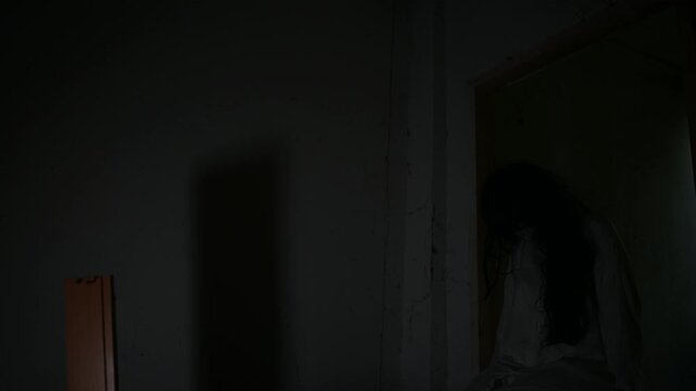 Dark horror scene of a mysterious figure in a white dress sitting alone in a dim abandoned room. Eerie shadows create a supernatural atmosphere for haunted house, paranormal and Halloween concepts.