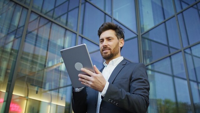 Handsome Caucasian businessman using tablet device near modern glass office building. Focused male scrolling on display and reading information outdoors during workday. Using touch screen.