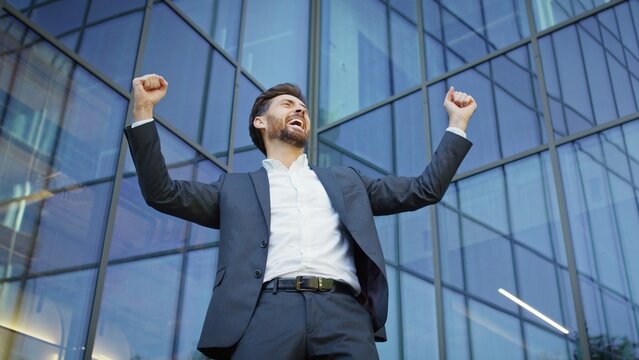 Successful businessman celebrating with arms raised in front of modern glass building