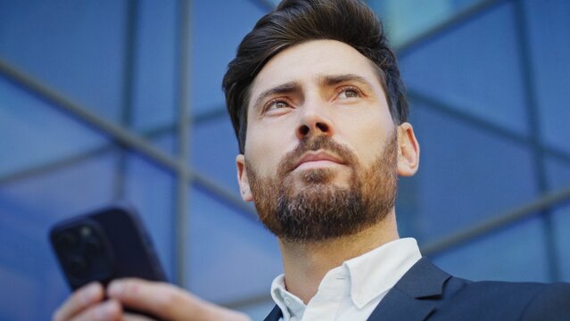 Attractive Caucasian male holding smartphone and thinking about best reply to email near glass office building. Focused businessman entering message on touch screen. Communicating with colleagues.