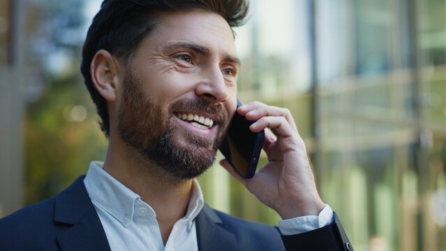 Smiling Caucasian businessman in formal suit having positive conversation on smartphone near glass office building. Male enjoying friendly talk outdoors during break. Smiling while communicating.