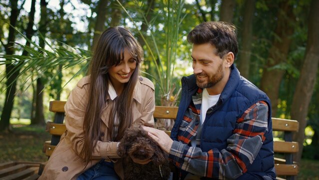 Caucasian couple sitting close on wooden bench smiling and petting small brown dog together. Man and woman enjoying peaceful romantic date in park surrounded by trees and soft daylight.