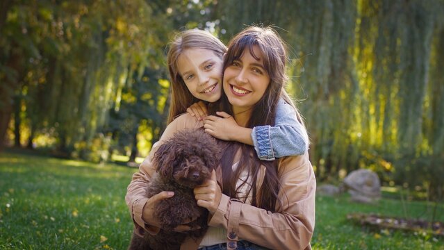 Smiling Caucasian woman and cheerful girl embracing while sitting on grass holding small brown dog. Mother and daughter spending relaxing afternoon outdoors in park surrounded by trees and sunlight.