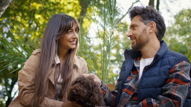 Caucasian man and woman sitting on bench smiling warmly at each other while holding small brown dog. Couple sharing loving moment outdoors surrounded by soft green trees and gentle sunlight.