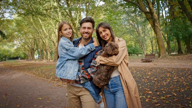 Smiling Caucasian family posing together outdoors in park. Man holding little girl while woman carrying small brown dog. Parents and child enjoying cheerful autumn day surrounded by fallen leaves.