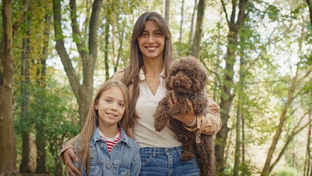 Smiling Caucasian woman standing with little girl holding small brown dog in arms. Mother and daughter enjoying peaceful walk together in forest park surrounded by trees and warm daylight.