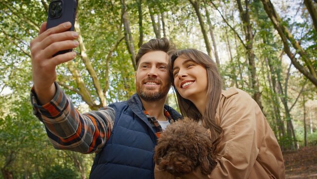 Caucasian couple taking selfie outdoors smiling warmly while woman holding small brown dog. Man and woman standing close enjoying joyful moment together surrounded by green trees and soft sunlight.
