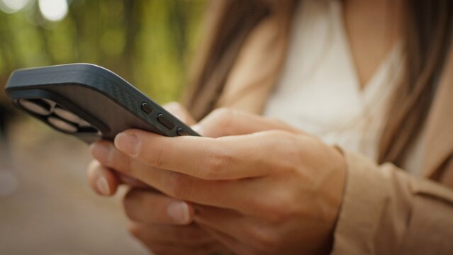 Close up of female hands holding smartphone outdoors typing on screen. Woman wearing beige coat using mobile device while standing in park with blurred trees. Remote communication concept.