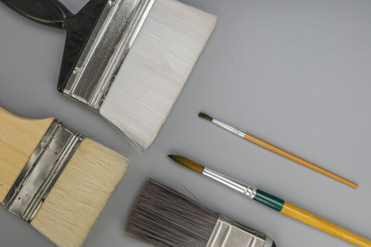 Flat lay composition of assorted wide wall painting brushes and small fine point watercolor artist brushes with wooden handles organized on a neutral gray background for creative art craft concept