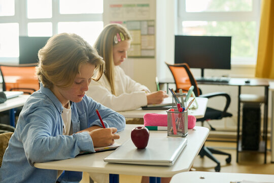 Caucasian kid writing in notebook at desk in classroom, classmate studying on background. Image represents primary school education, suitable for academic marketing campaigns