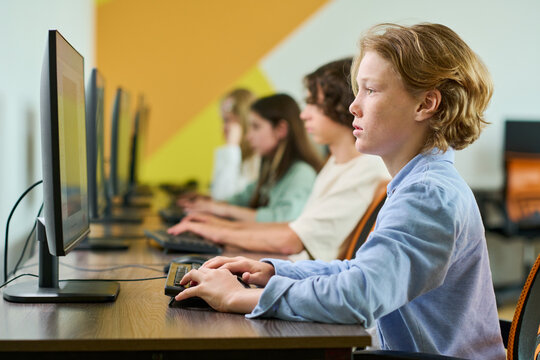 Caucasian teenager boy using computer in classroom with classmates. Image represents digital literacy and STEM education, suitable for school marketing materials, eye level shot