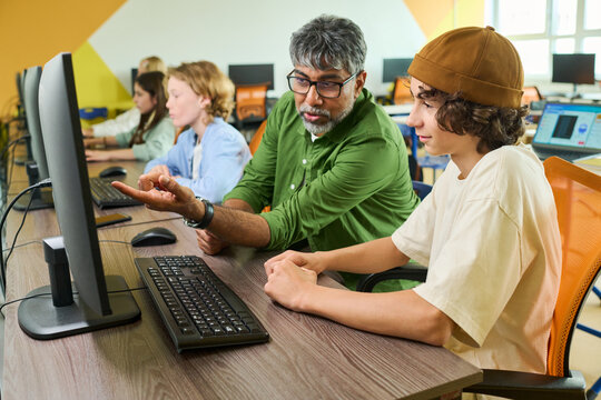 Middle aged Indian male teacher explaining computer programming to Caucasian teenager boy in classroom. Image represents STEM education and digital literacy training for youth