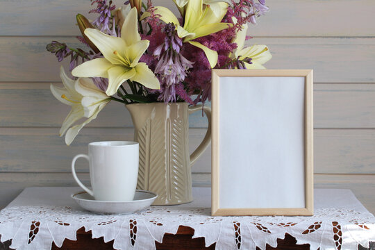 A rural mockup with an empty frame, a white mug, and a bouquet of garden lilies, astilbe, and hostas in a jug on the table. Empty space.