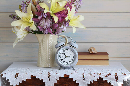 A rural still life with a bouquet of garden lilies, astilbe, and hostas in a jug on a table with a retro alarm clock.