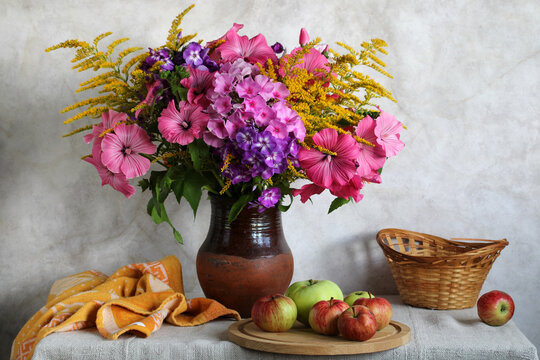 summer still life with pink lavatera, phlox, and yellow goldenrod in a clay jug on the kitchen table. Garden flowers and apples.