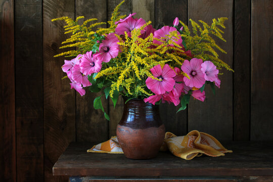Pink lavatera and yellow goldenrod in a clay jug on a wooden table; a still life with flowers against a dark background.