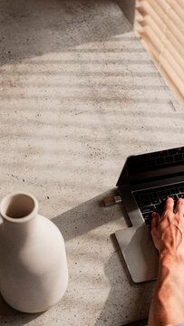 Top view of a person typing on a laptop keyboard at a minimalist concrete desk illuminated by natural sunlight through window blinds