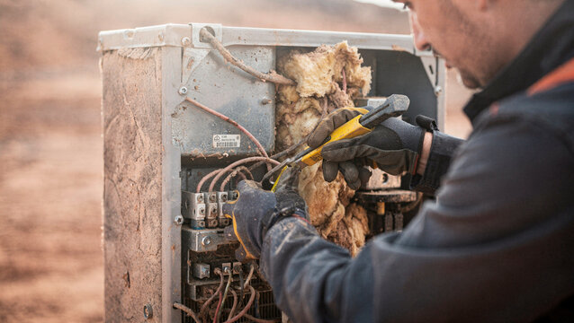 An electrician in protective gloves repairs electrical wiring inside an outdoor control box using a utility knife