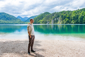 Fototapeta premium Bald young man wearing glasses, white hoodie, plaid trousers, Travel tourism stylish clothing choices, pebble beach, turquoise lake, mountains, forest trendy attire, summer vacation, fashion portrait