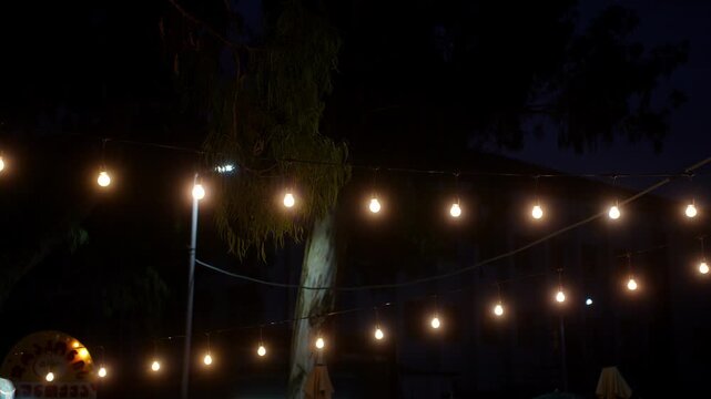 Outdoor string lights glowing at night close up, warm light bulbs garland hanging in summer cafe terrace, cozy party illumination and backyard festive decor background.