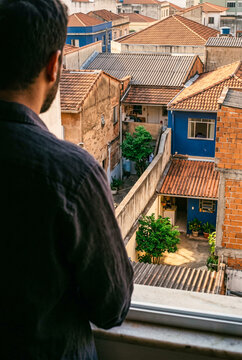 A man stands at a window looking out at a traditional residential neighborhood with tiled roofs and colorful houses