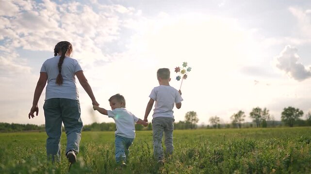 Girl walks with two boys across field. Toddler holds sibling hand walking slowly in nature. Child steps forward with girl and boy holding hands. Outdoor walk in field with toddler and sibling.