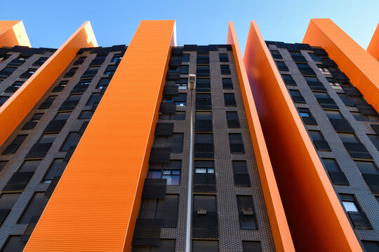 Low-angle view of a modern high-rise building with dramatic orange vertical fins alternating with dark brick facade sections, rising against a clear blue sky.