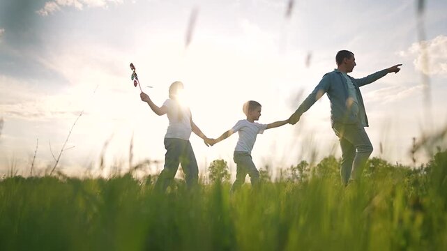 Father walks ahead in grassy field. Boy walks between father and girl. Girl waves toy and smiles. Father holds child hand. Grass and nature surround boy girl holding father hand while walking forward